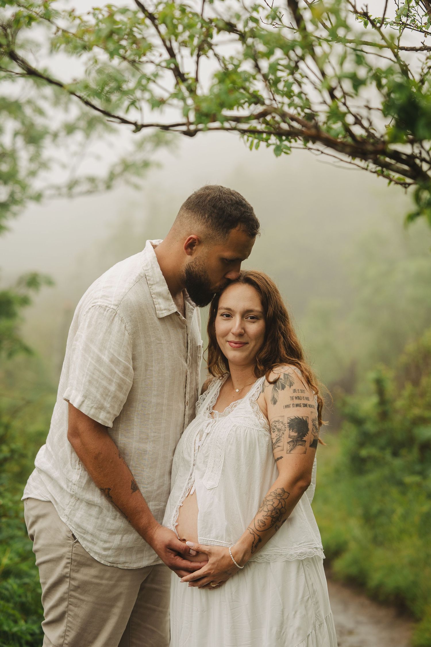 Couple in white outfits embracing outdoors in misty green forest, pregnant woman smiling, man kissing her head tenderly.