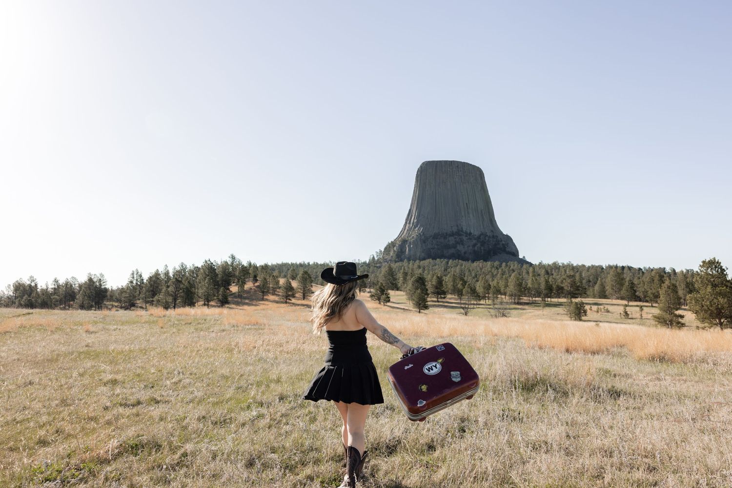 Woman in black dress holding vintage suitcase walks toward Devils Tower, Wyoming landmark, in open field.