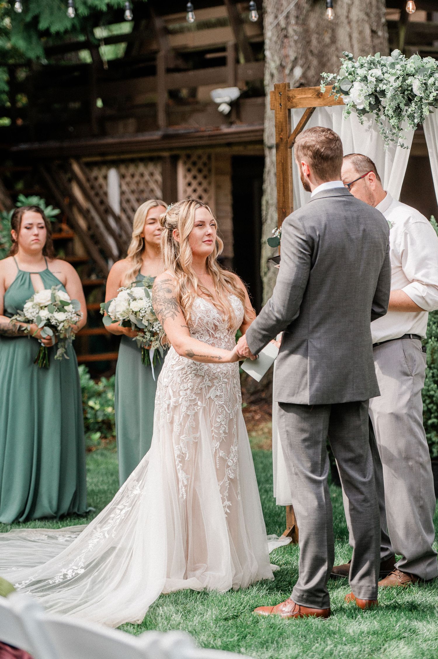 Bride in lace gown holds hands with groom at outdoor wedding ceremony, bridesmaids in sage green dresses stand behind.