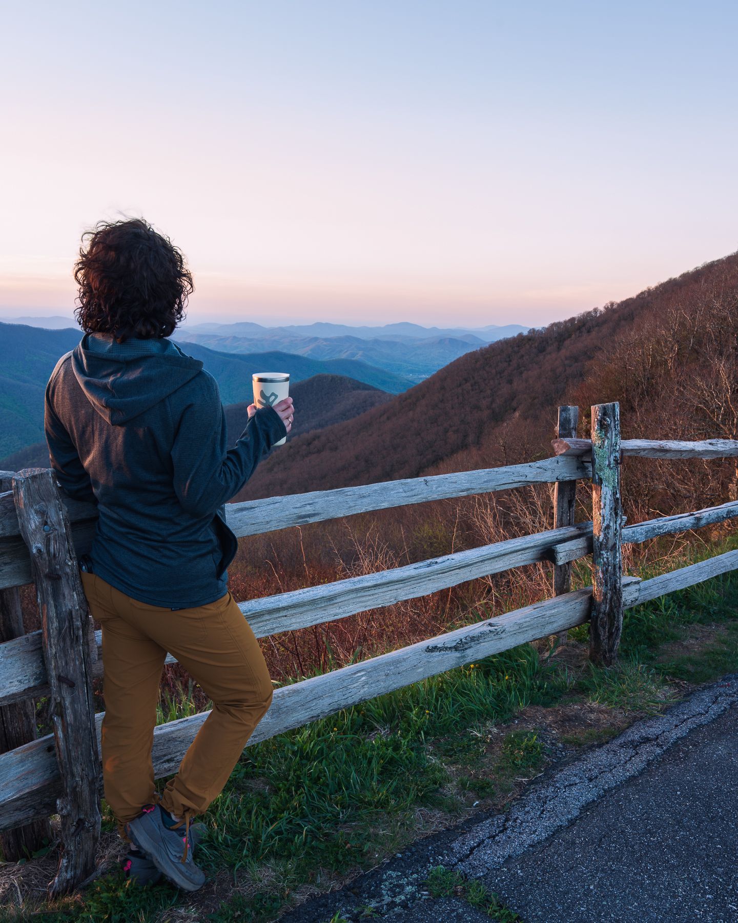 A person in a blue jacket holds a cup while leaning on a wooden fence overlooking misty mountain ranges at sunset.