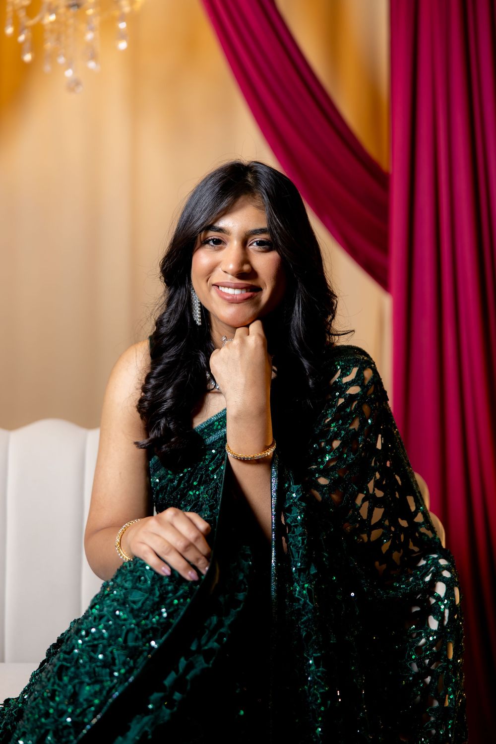 A smiling woman in an elegant emerald green sequined saree poses gracefully against a gold and crimson draped backdrop.