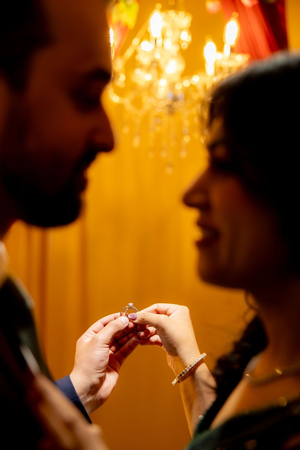 Romantic couple sharing an intimate moment while holding an engagement ring, with warm chandelier lighting in the background.