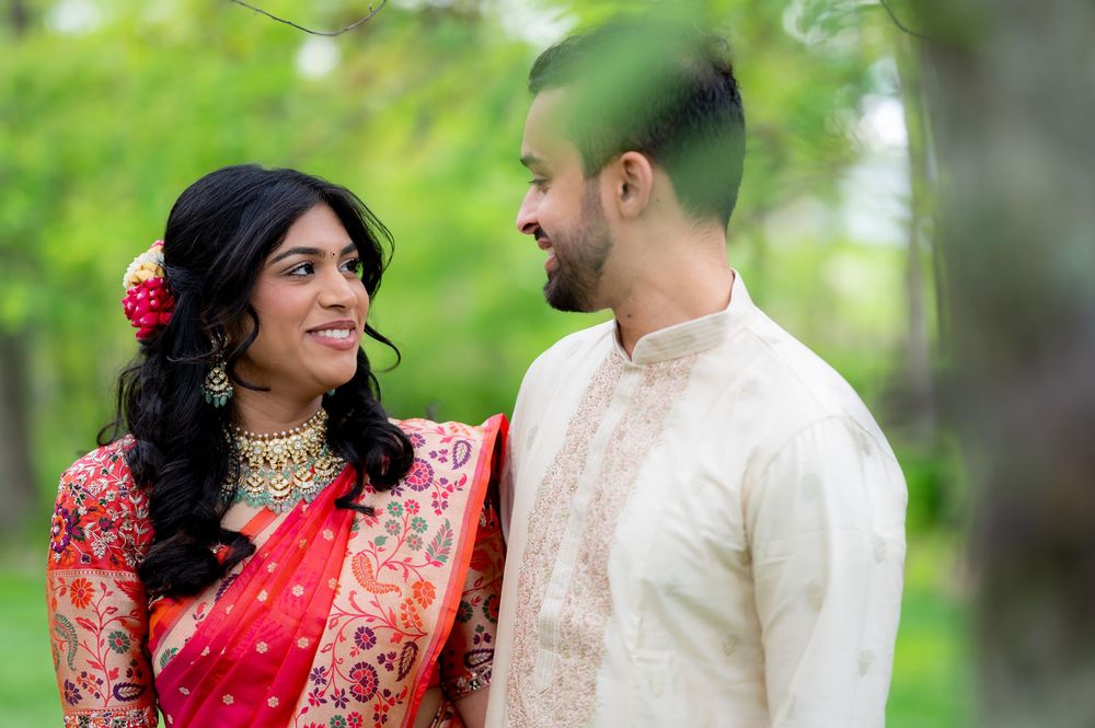 Couple in traditional Indian attire smiling at each other outdoors, woman in red saree with floral hair accessory.