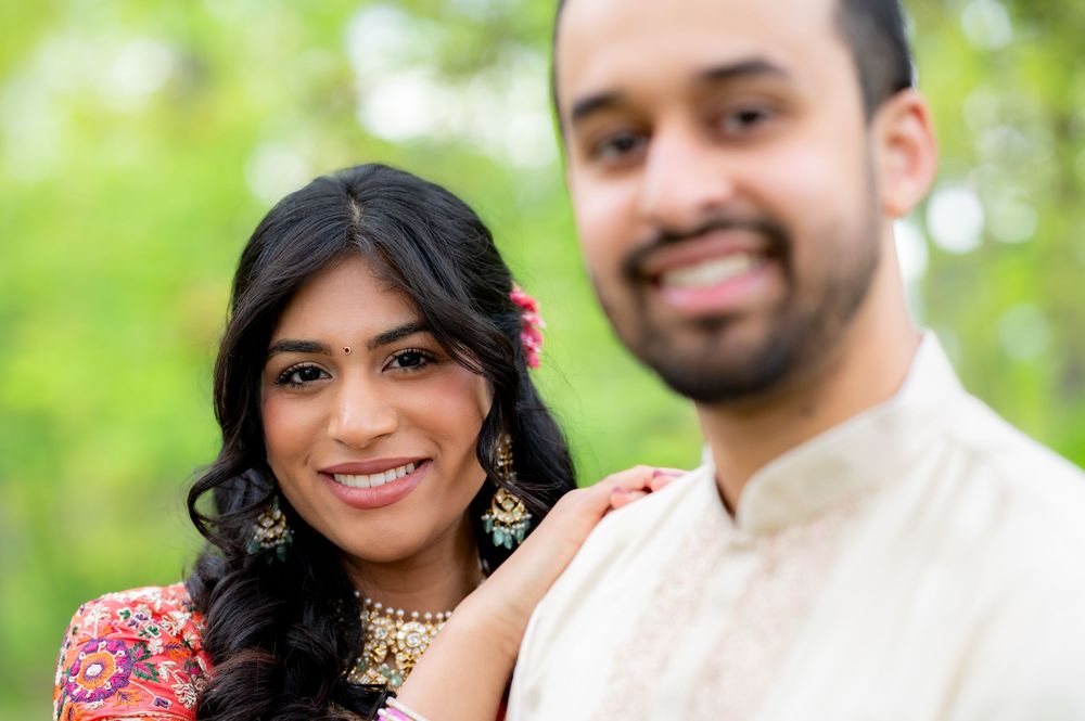 Indian couple in traditional attire smiling outdoors, woman in floral dress with gold jewelry, man in cream kurta.