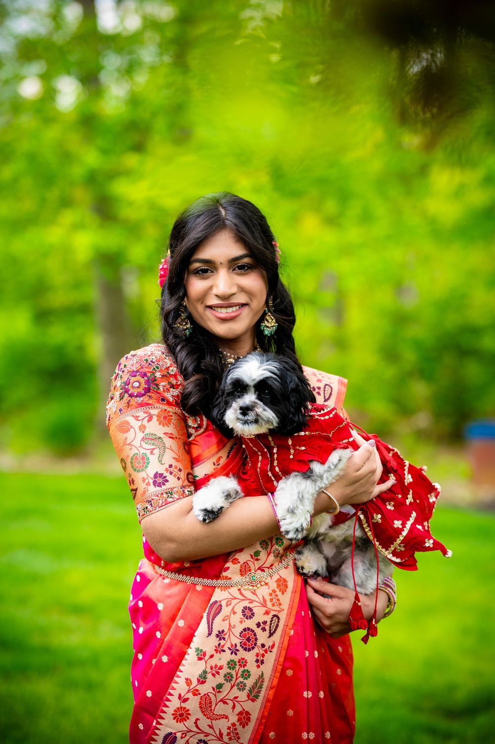 Woman in red saree smiling and holding a small Shih Tzu dog dressed in red, posing outdoors.