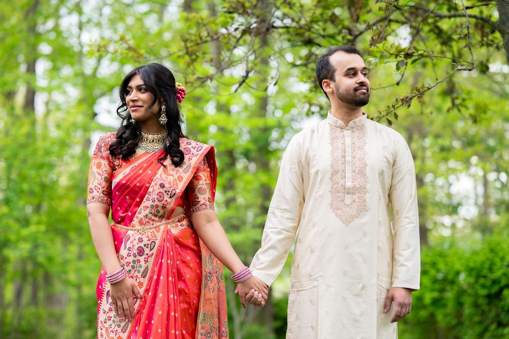 Indian couple in traditional wedding attire holding hands outdoors, bride in red saree, groom in cream kurta.