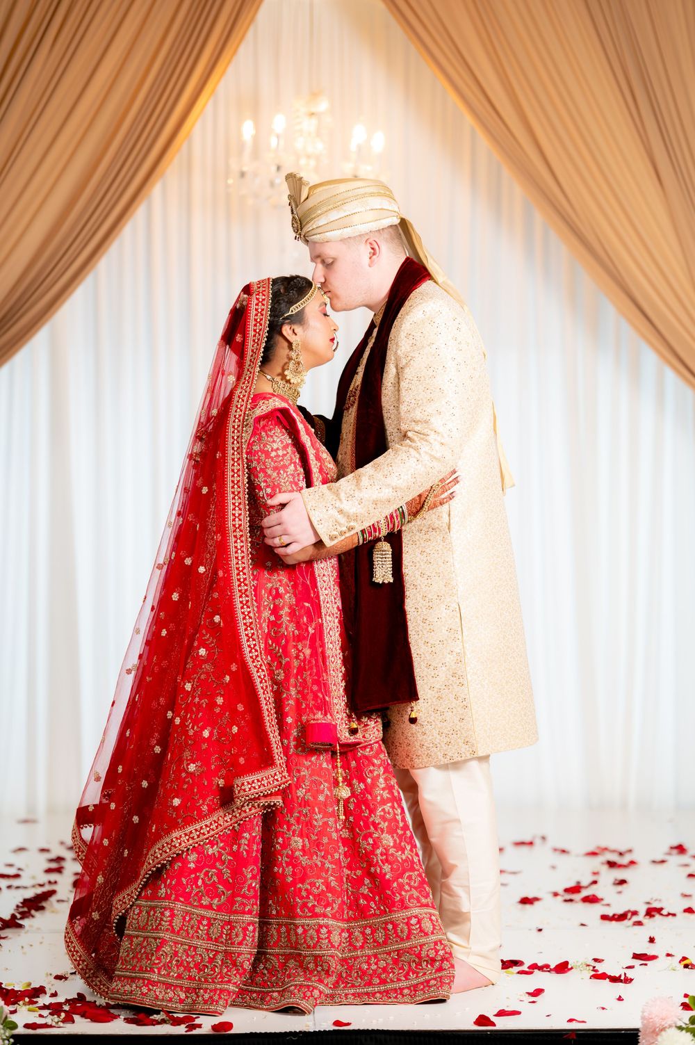 Indian bride in red lehenga and groom in cream sherwani share a kiss at their traditional Hindu wedding ceremony.