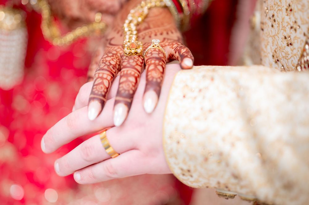 Close-up of bride's intricate mehndi henna hands held by groom wearing a gold wedding band at Indian wedding.