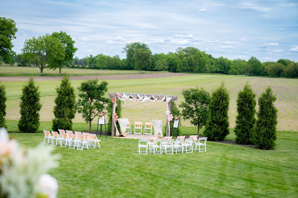Outdoor wedding ceremony setup with white chairs and floral arch on lush green lawn with scenic farm views.