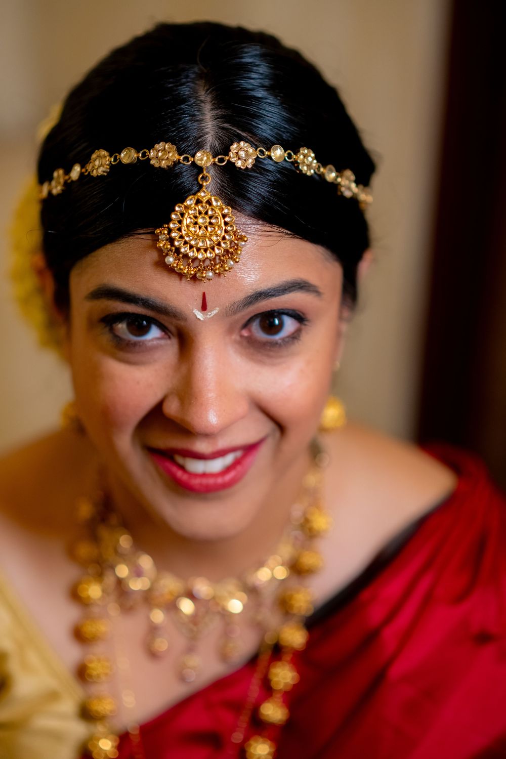 Indian bride smiling in red saree with gold maang tikka, necklace, and traditional bridal jewelry.