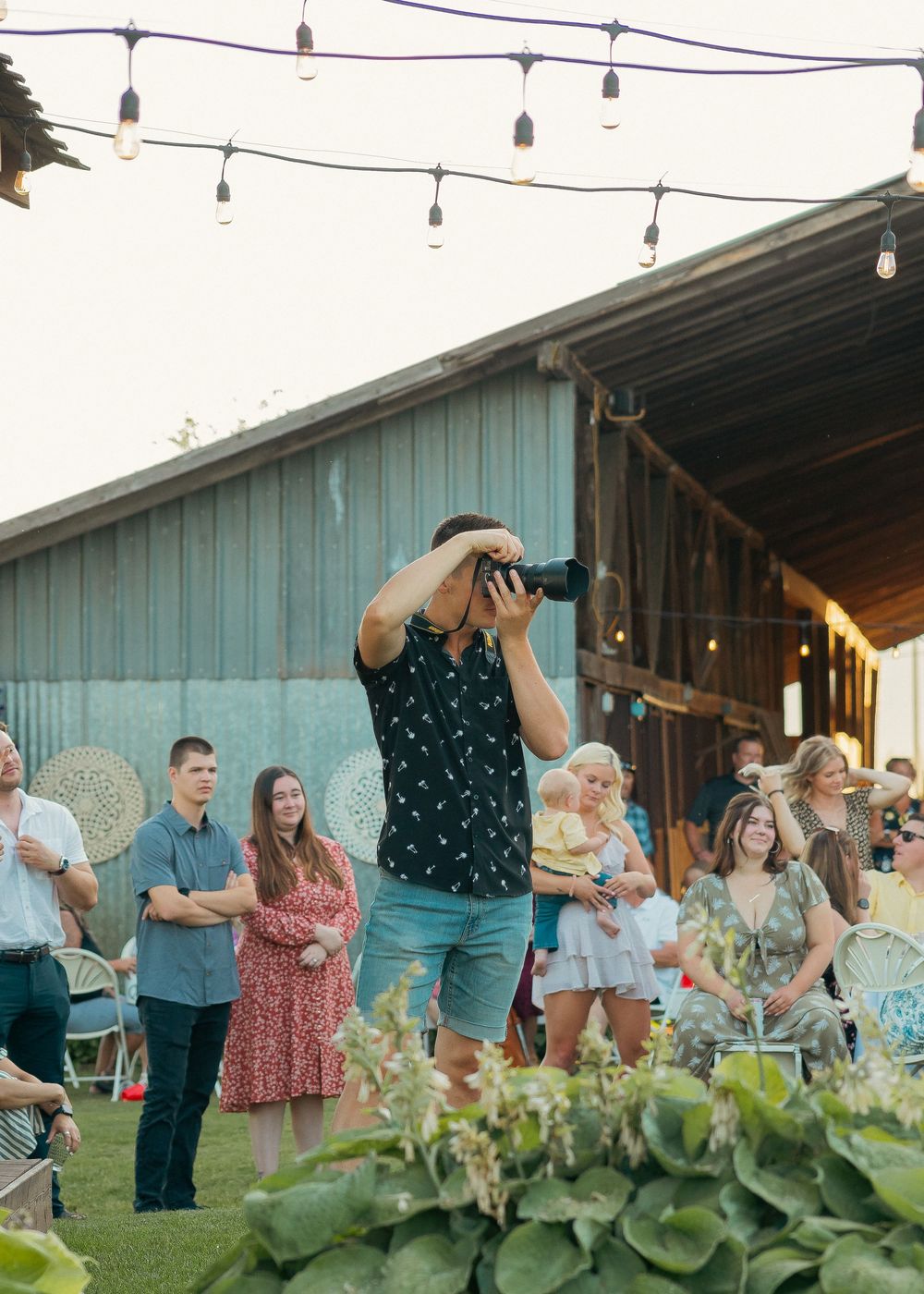 Photographer capturing moments at an outdoor barn wedding reception with string lights and guests gathered around.