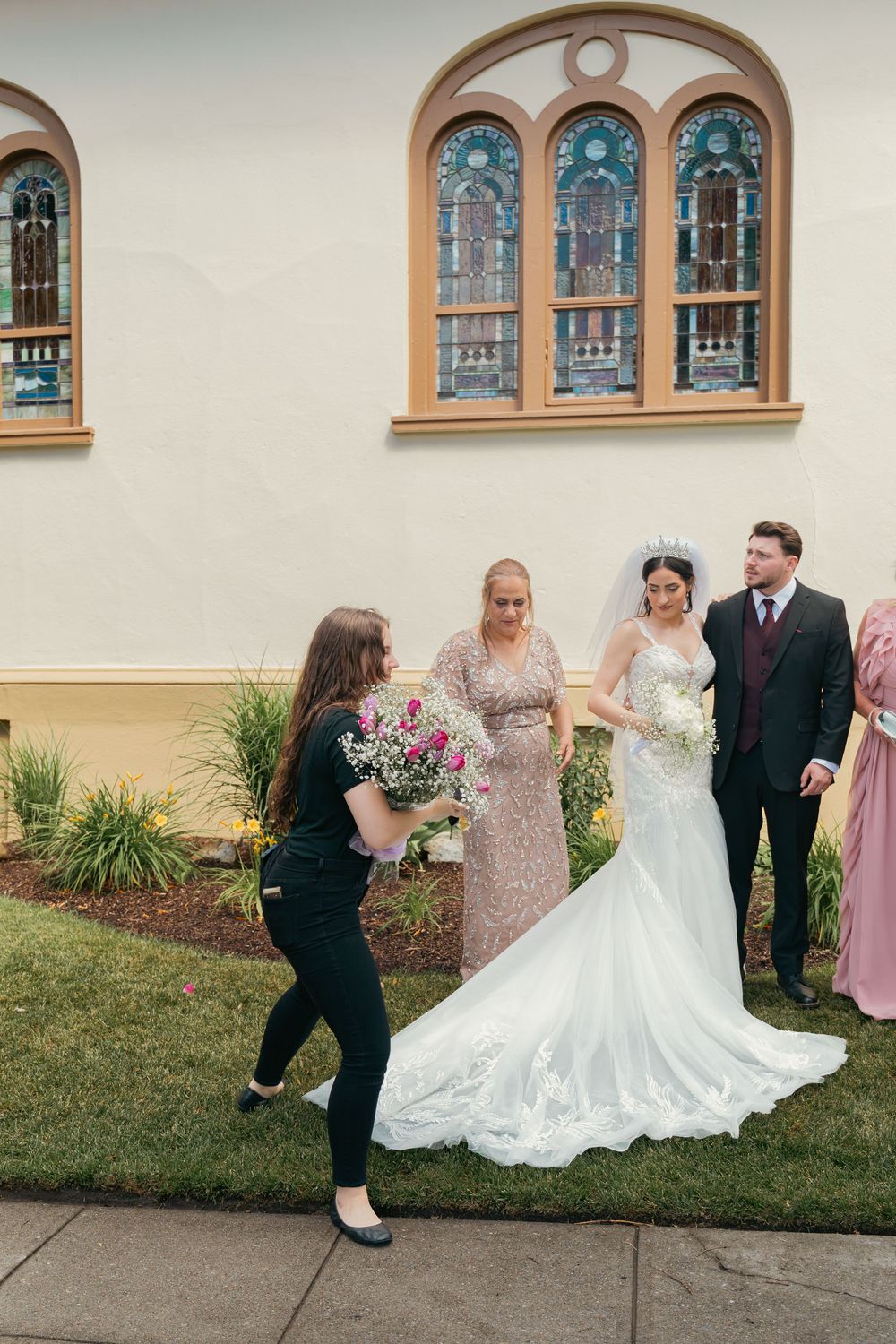 Wedding coordinator hands bouquet to bride in lace gown outside a church with stained glass windows.