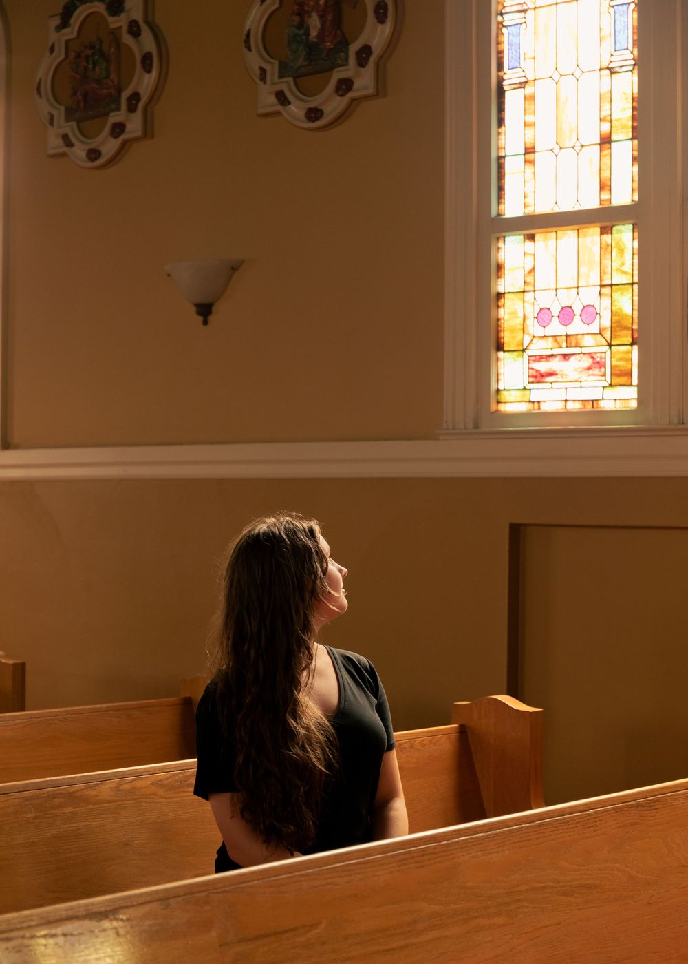 Woman sitting alone in church pew gazing at colorful stained glass window in sunlit chapel.