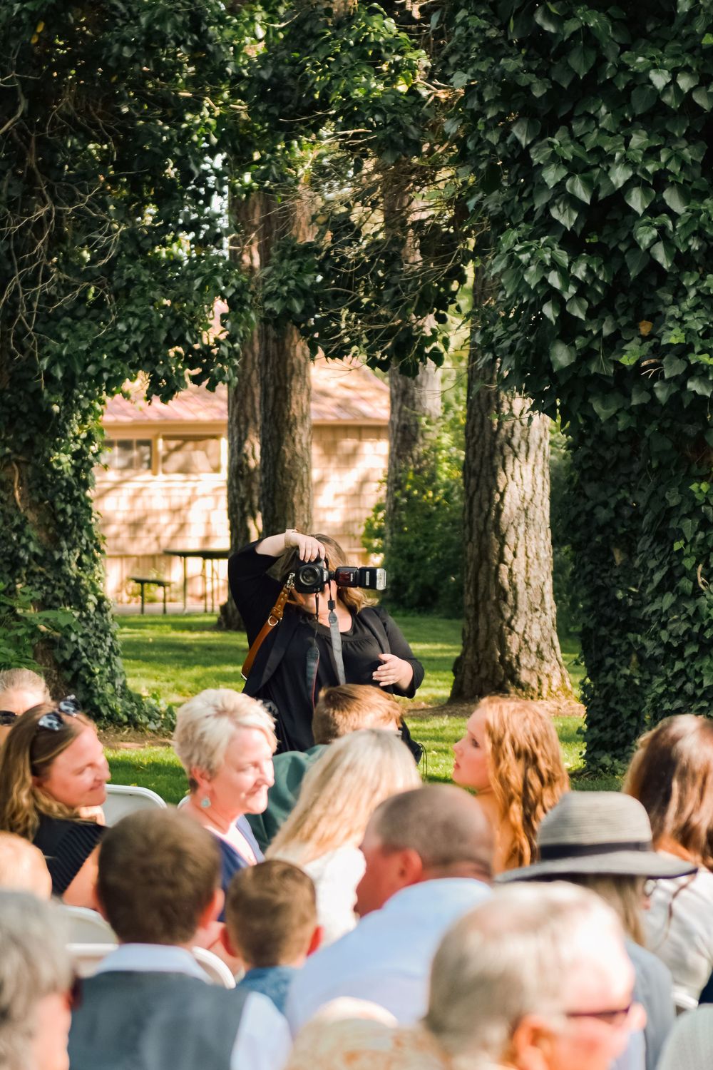 Photographer capturing moments among wedding guests seated outdoors under lush green tree canopy.