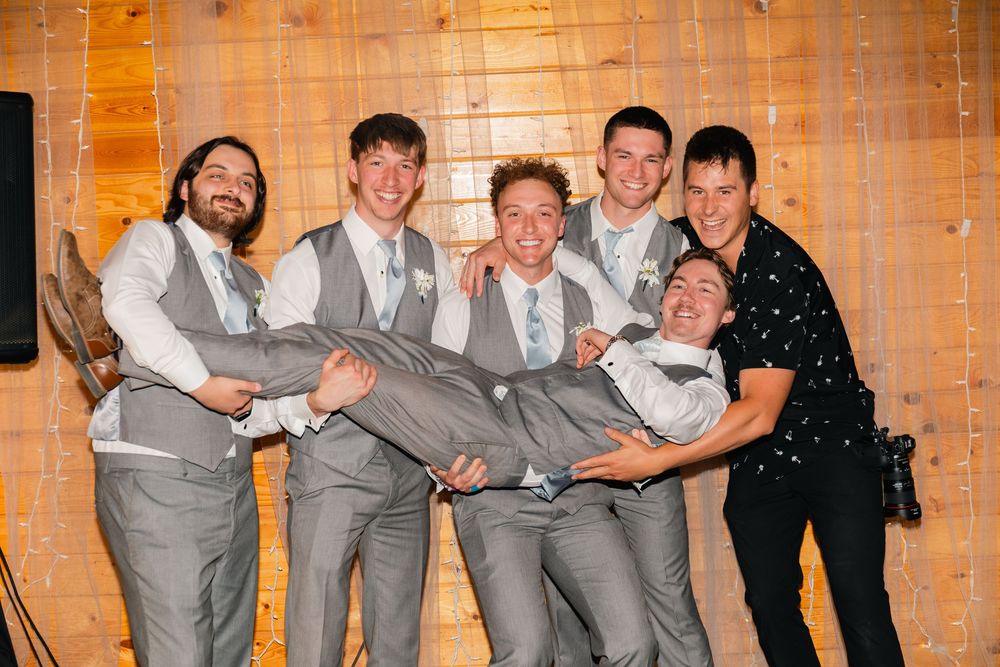 Groomsmen in gray vests lifting groom while wedding coordinator poses with group indoors.
