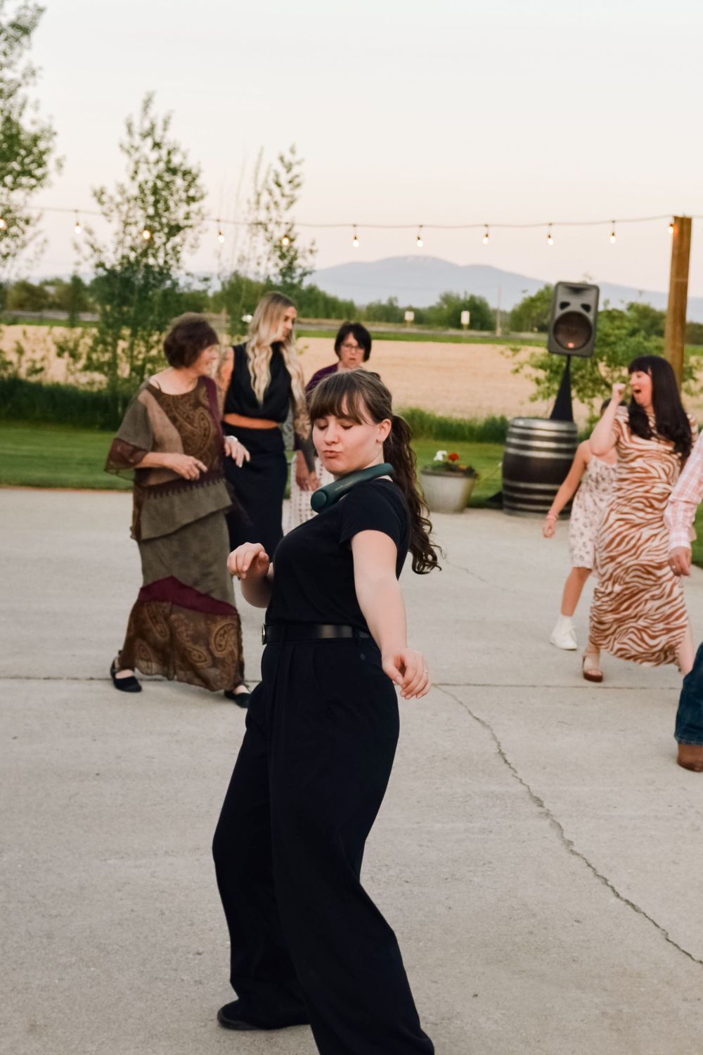 Woman in black outfit dancing outdoors at an evening event with string lights and guests.