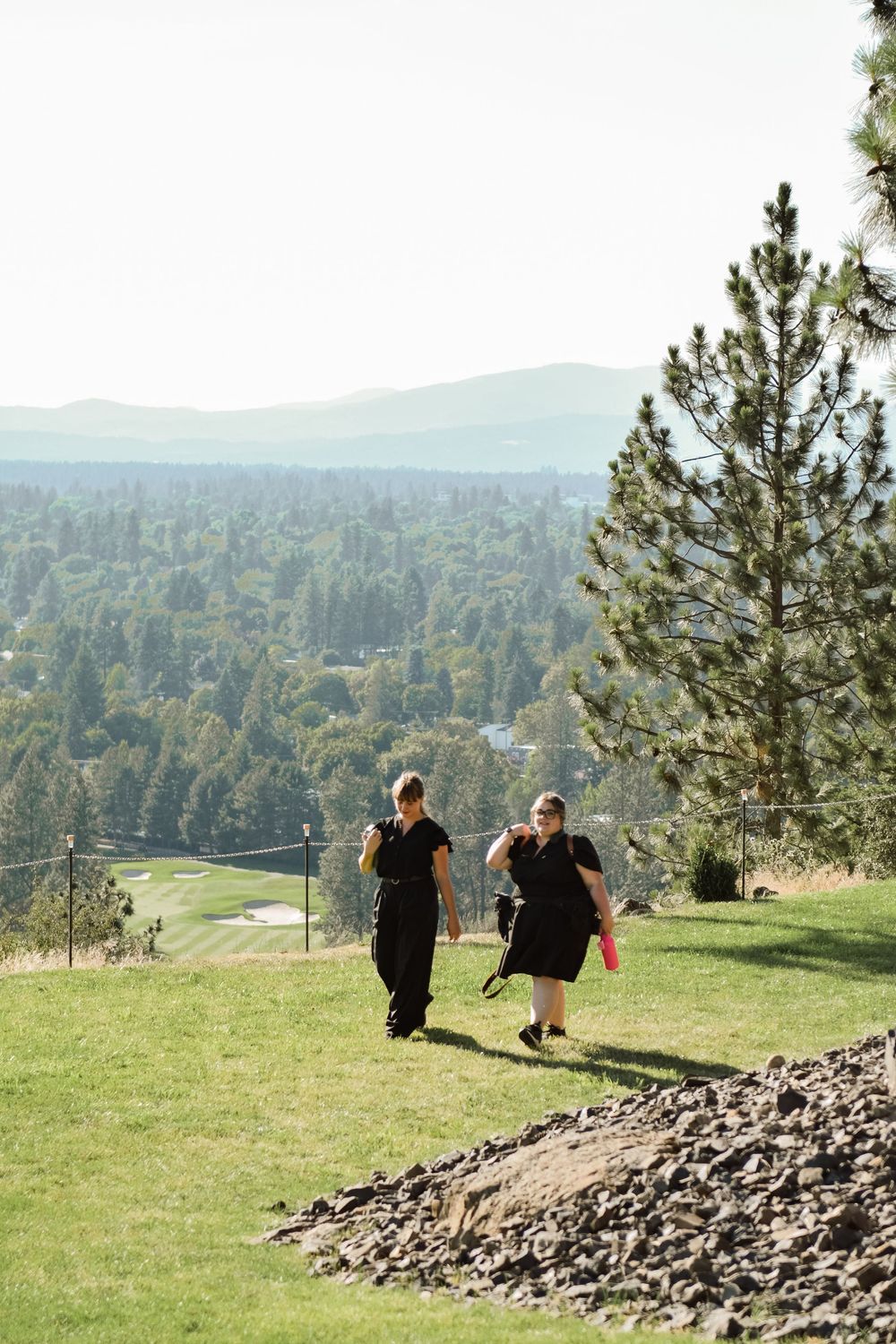 Two women in black walking on grassy hillside with pine trees and mountain views.