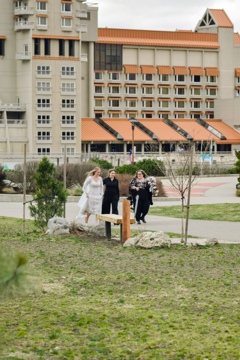 Bride in white gown walks with two guests near a large hotel building in an outdoor park setting.
