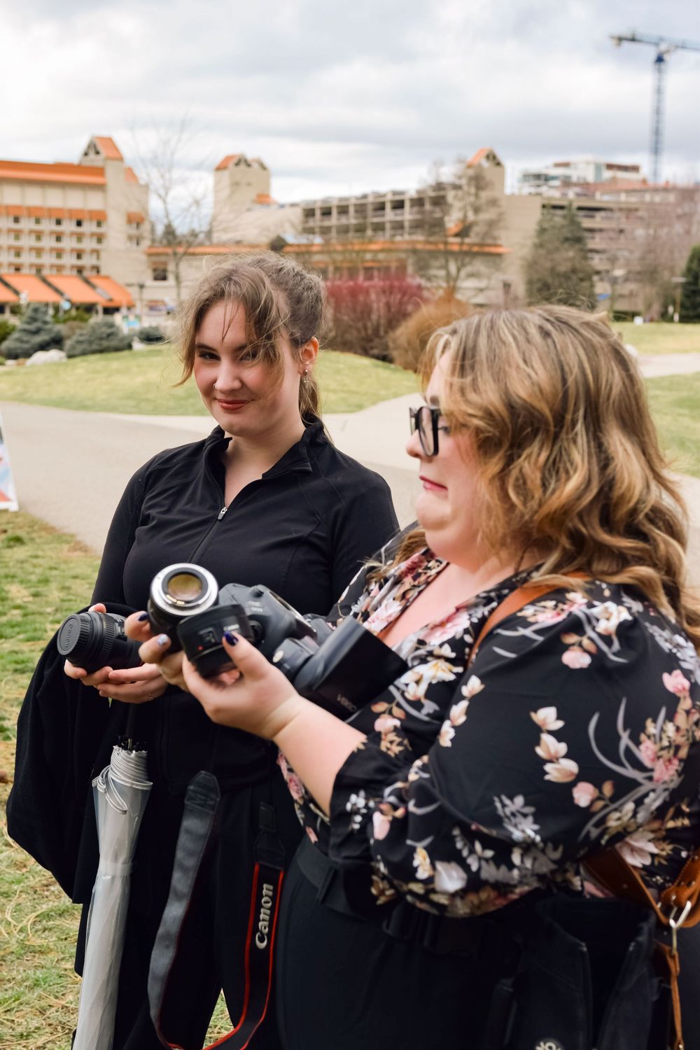 Two women photographers reviewing images on a Canon camera outdoors near a park with buildings in the background.