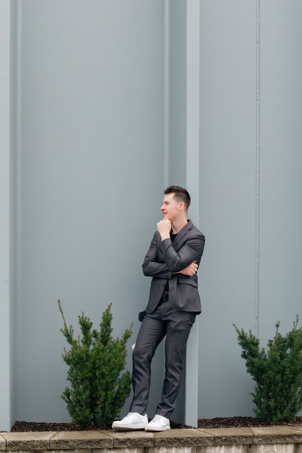 Stylish young man in gray suit posing thoughtfully against gray wall with small evergreen shrubs outdoors.