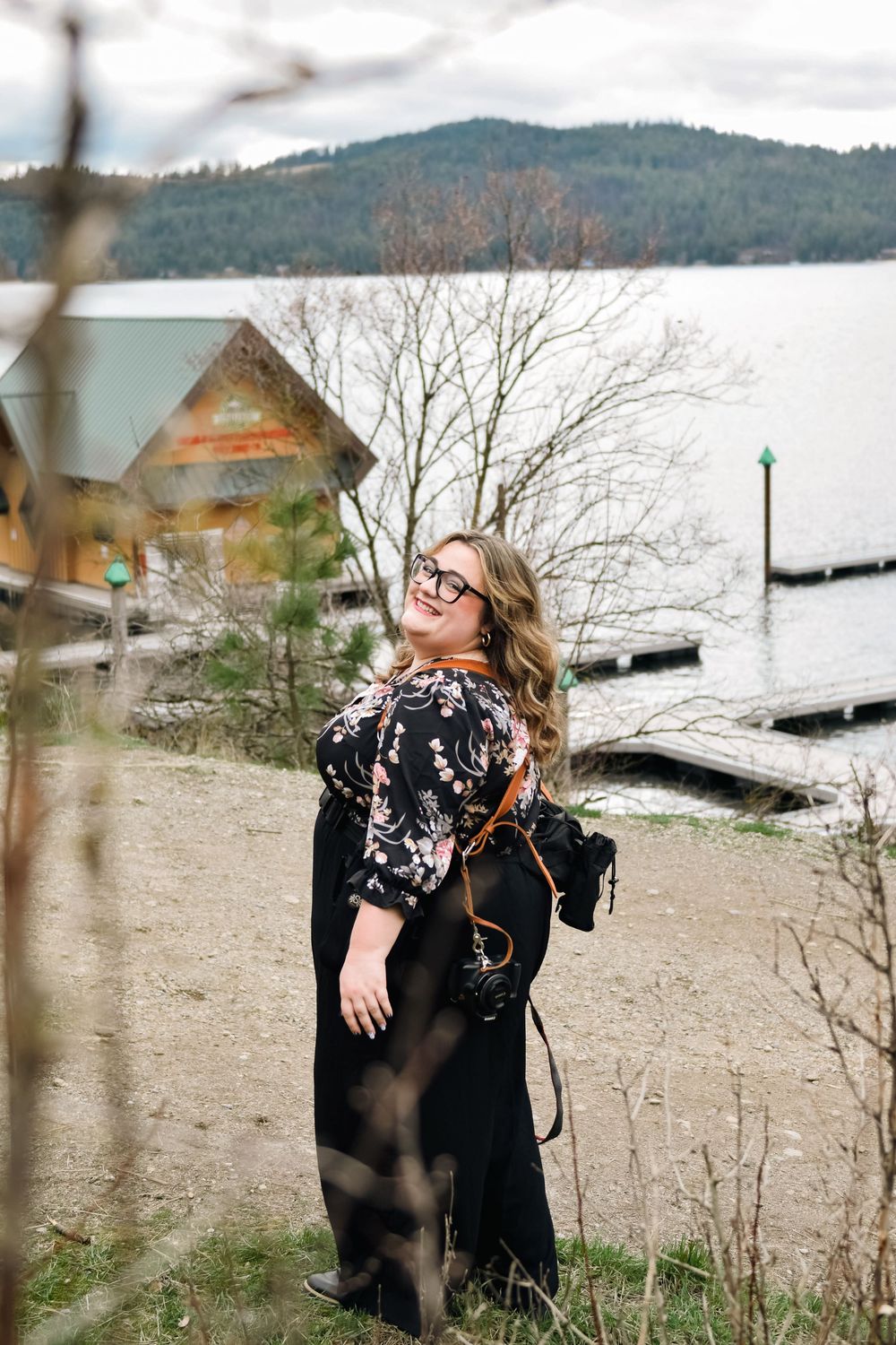 Female wedding photographer with camera and glasses posing near lakeside dock with mountain backdrop.