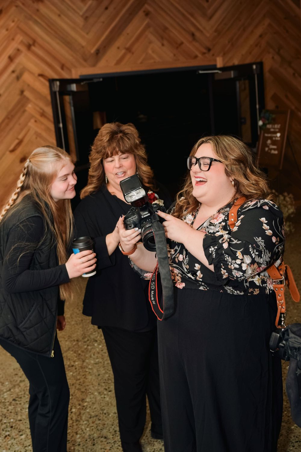 Three women reviewing wedding photos on camera with flash at rustic barn venue.