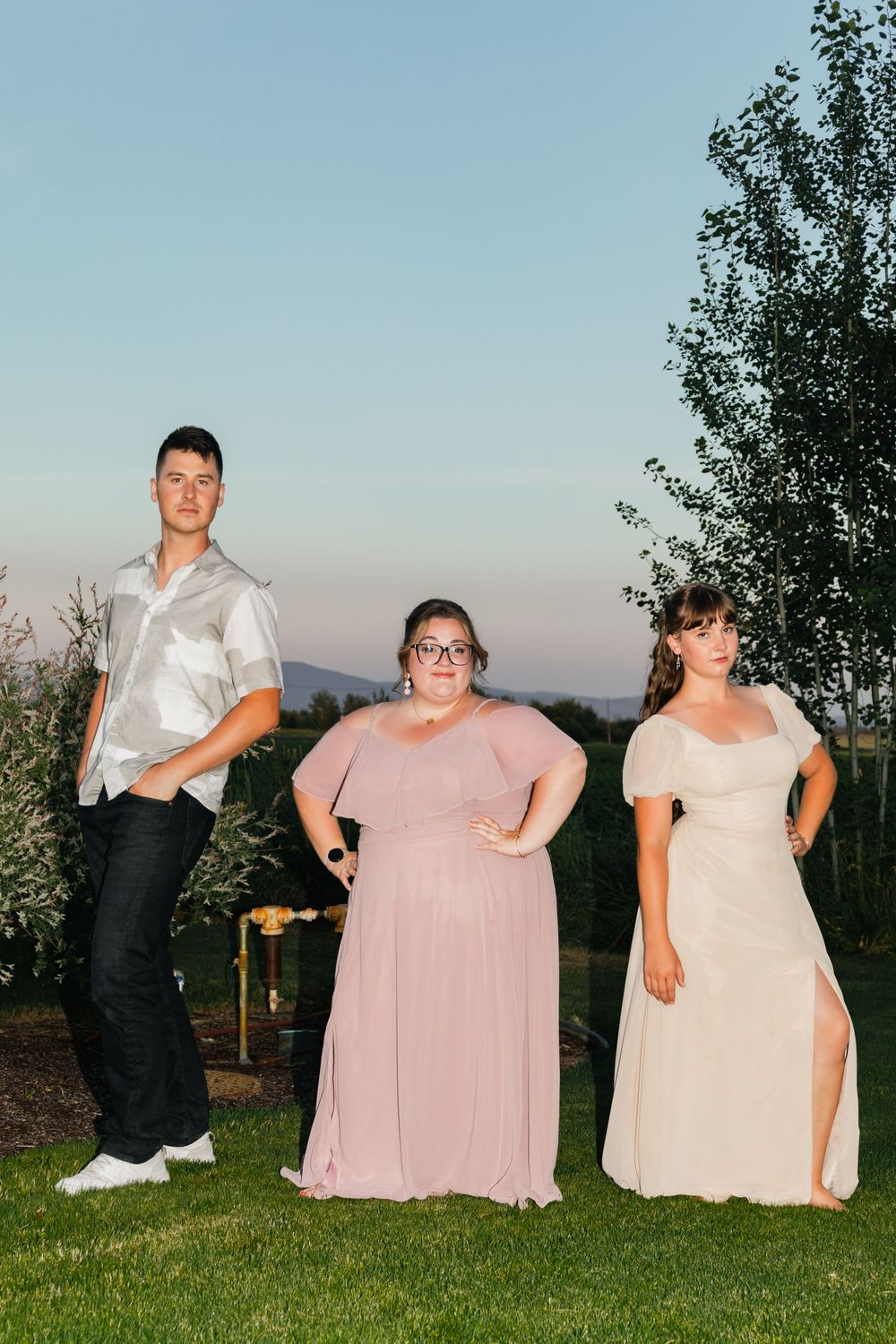 Three people posing outdoors at dusk, wearing formal attire including pink and cream gowns.