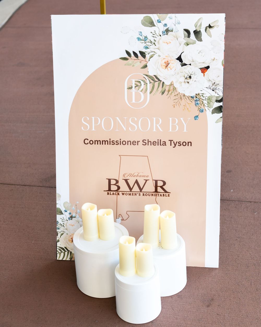 Sponsor sign for Commissioner Sheila Tyson at Black Women's Roundtable event with floral decor and white candles.