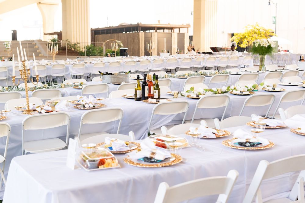 Elegant outdoor wedding reception with long white tables, gold chargers, floral centerpieces, and white folding chairs set under a tent.