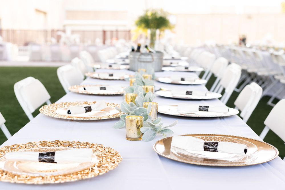 Elegant outdoor wedding reception table set with gold charger plates, white napkins, and greenery centerpieces on a white tablecloth.