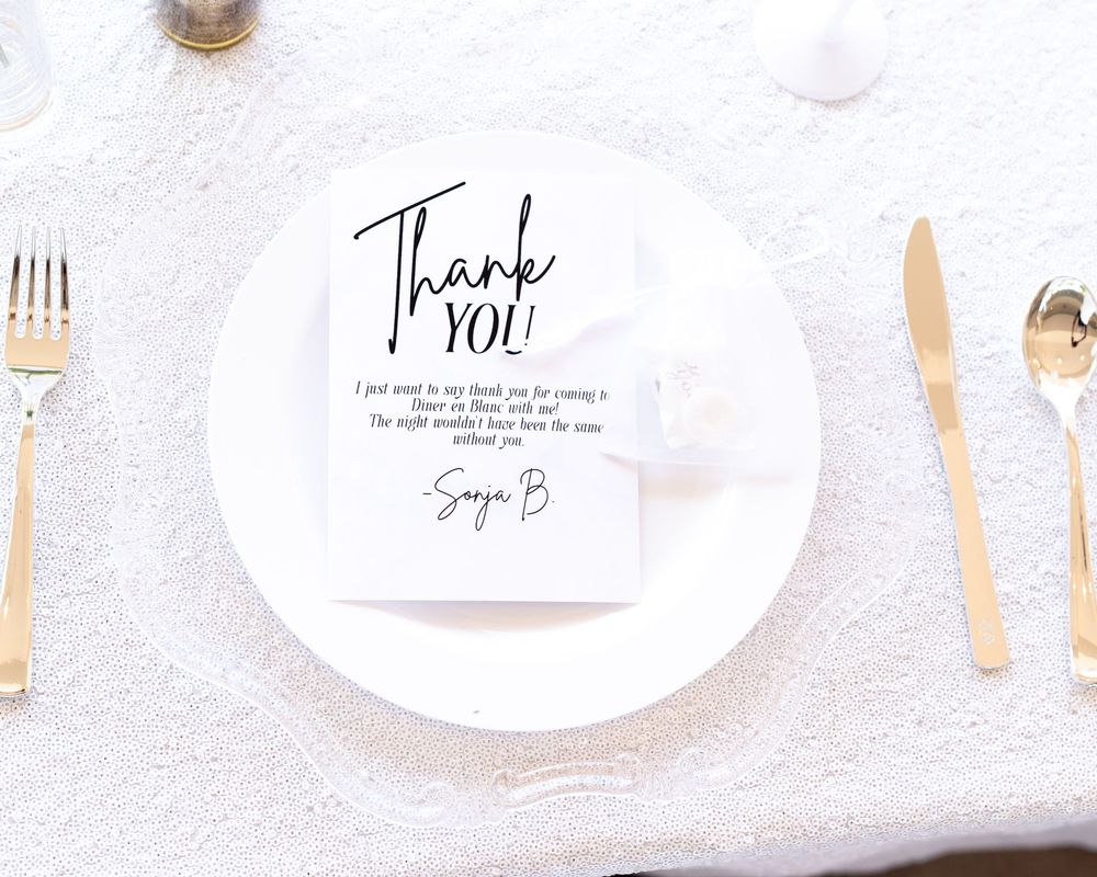 Elegant white place setting with gold cutlery and a personalized thank you card on a textured white tablecloth.
