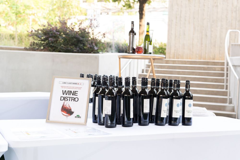 Wine distro table displaying rows of red wine bottles at an outdoor event with a decorative sign.