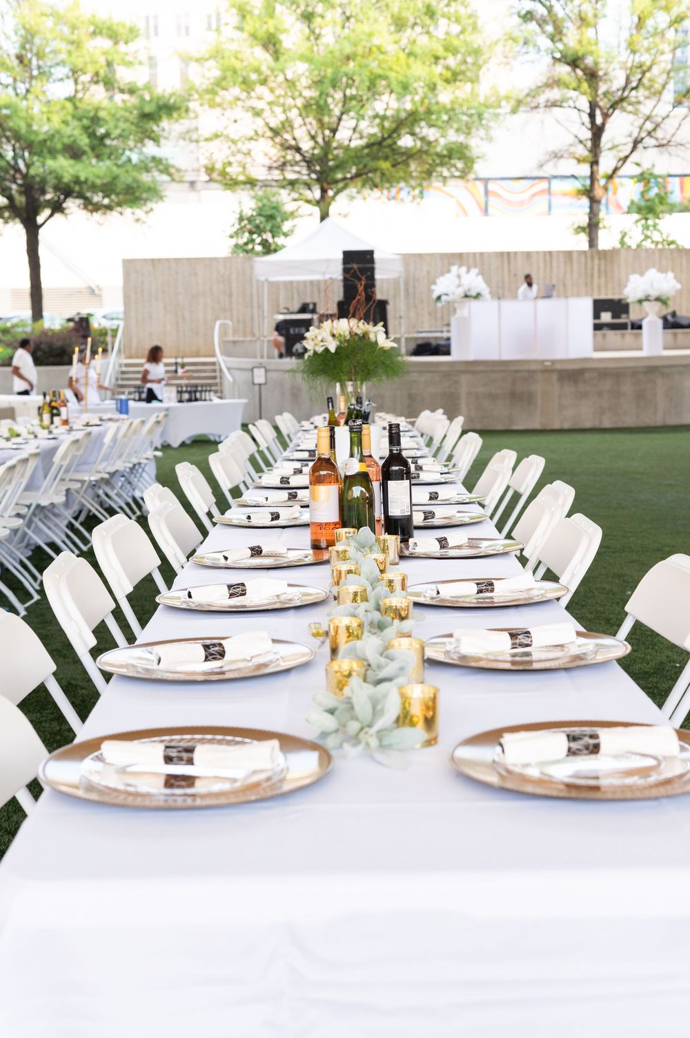Elegant outdoor wedding reception with long white tables set with gold chargers, wine bottles, and floral centerpieces.
