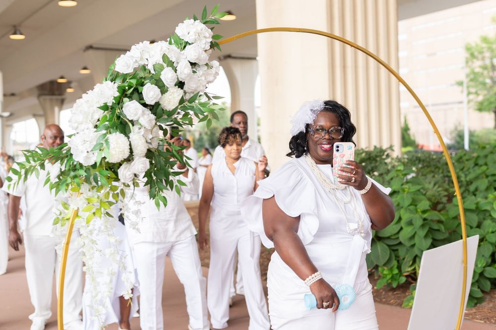 Guests dressed in white attire walk under a gold circular arch adorned with white flowers at an outdoor event.
