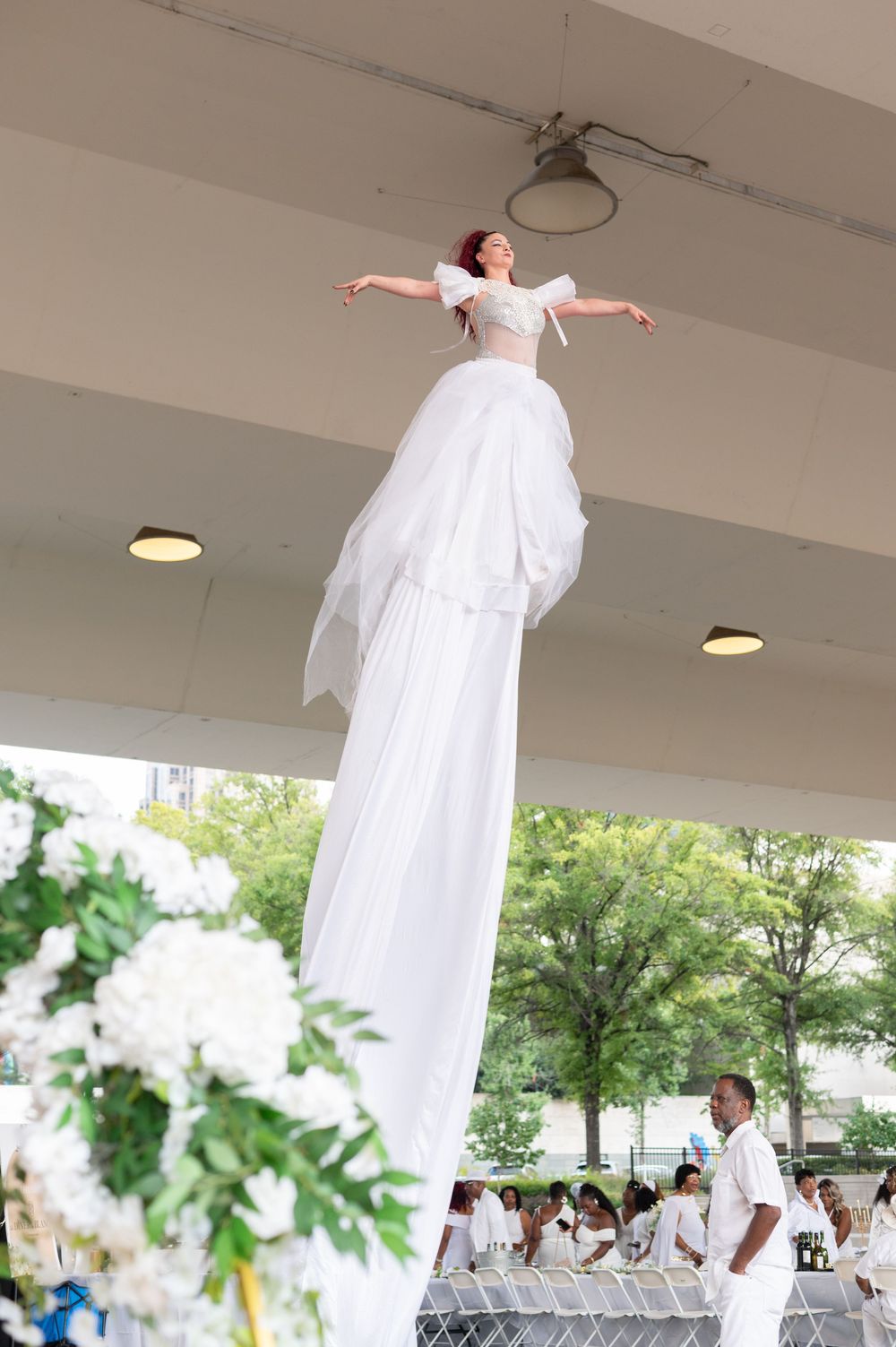 Performer on stilts in elegant white gown with arms outstretched at an outdoor event with white floral decor.