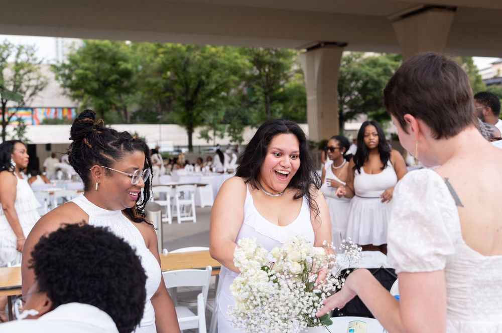 Women in white dresses laughing and socializing at an outdoor event, one holding a bouquet of baby's breath flowers.