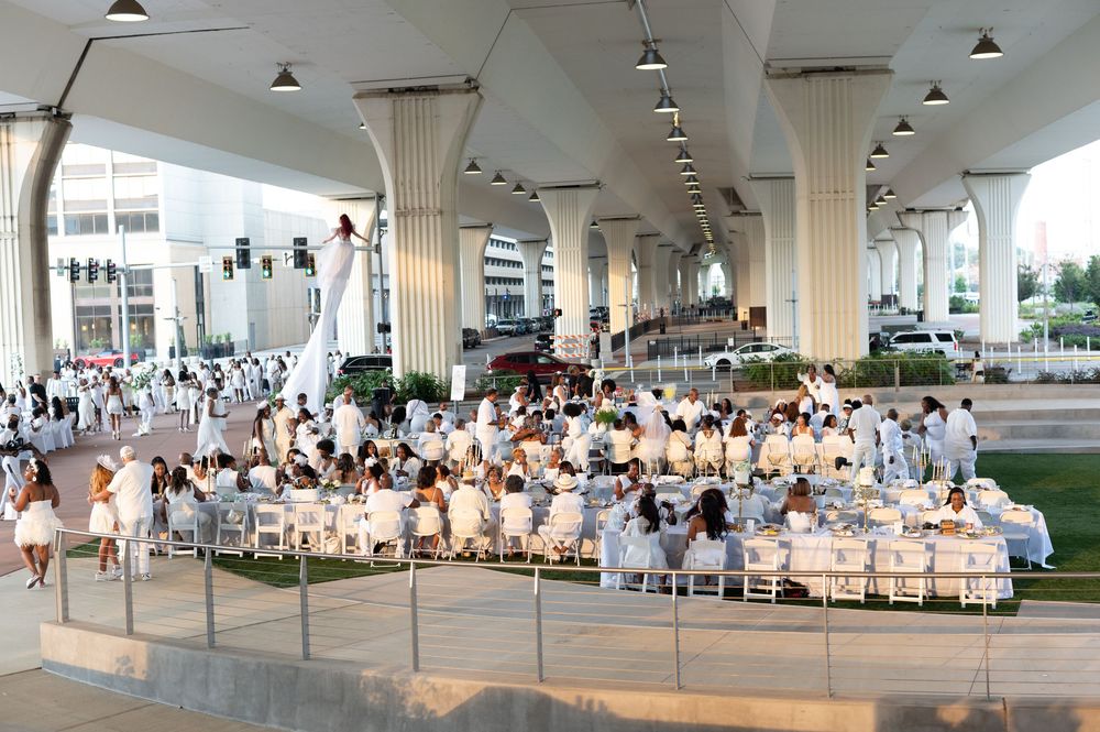 Large outdoor white-themed dining event held under elevated structure with guests seated at decorated tables.