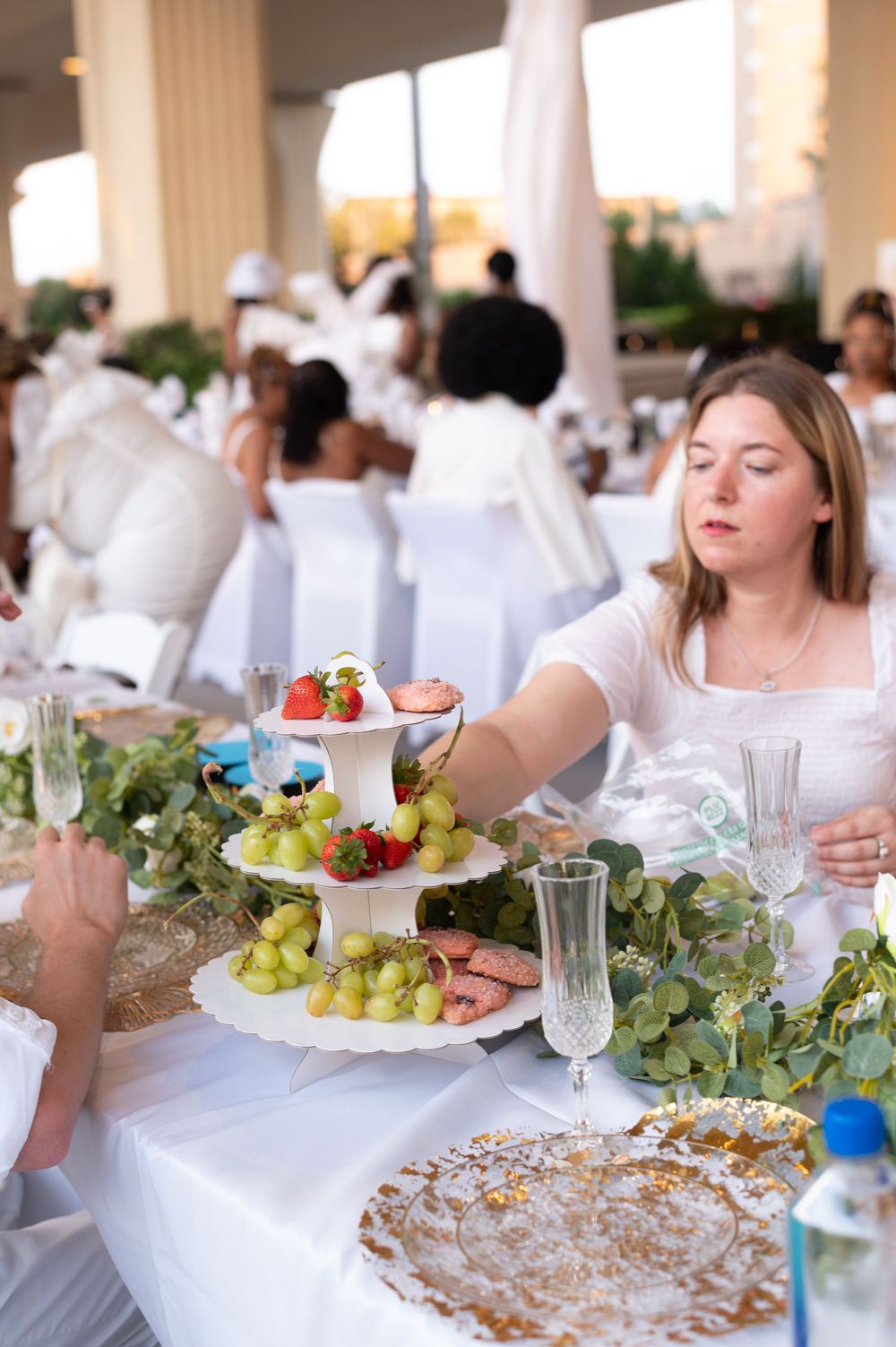 Woman in white dress reaches for tiered fruit display at elegant all-white outdoor dining event.