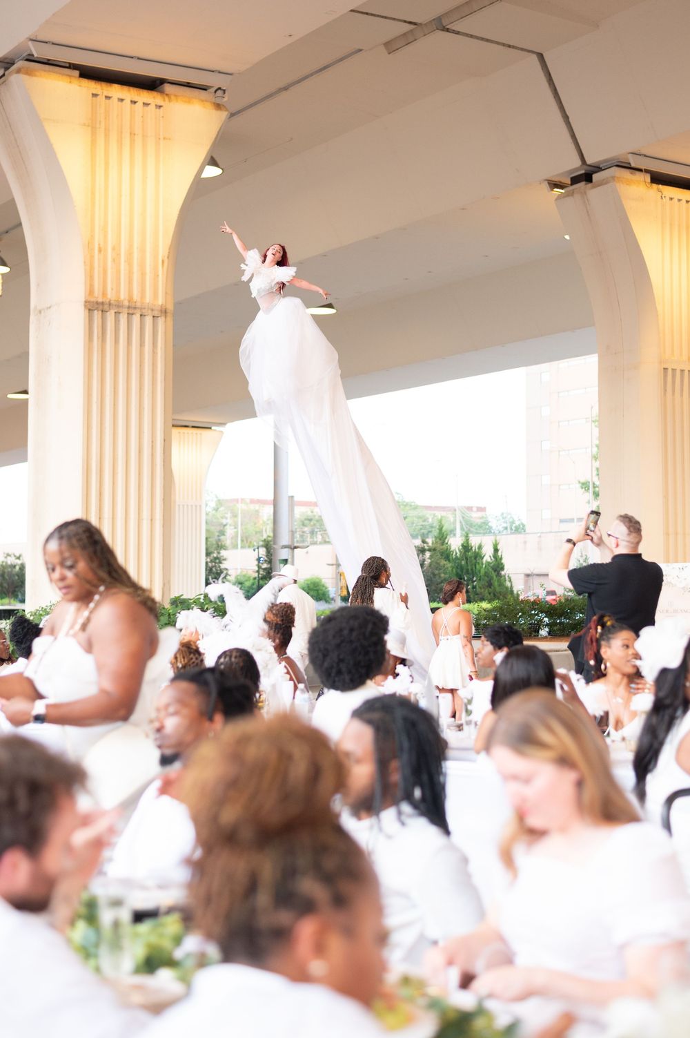 Elegant all-white outdoor dinner event with a performer in a white gown elevated above seated guests under arched columns.
