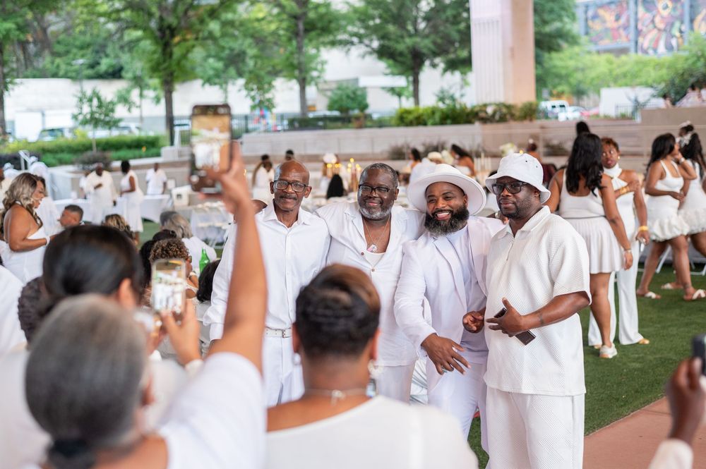 Group of elegantly dressed people in white attire posing for photos at an outdoor all-white party event.