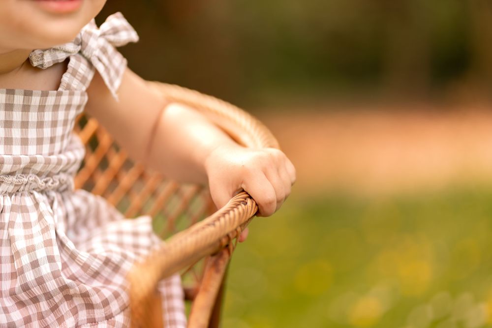 Young girl in a brown gingham dress sitting in a wicker chair outdoors in a garden setting.