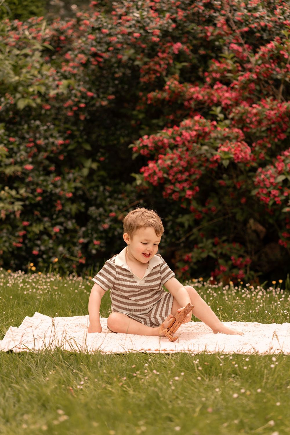 Young boy sitting on blanket in garden with pink flowering bushes, wearing striped romper, playing outdoors.