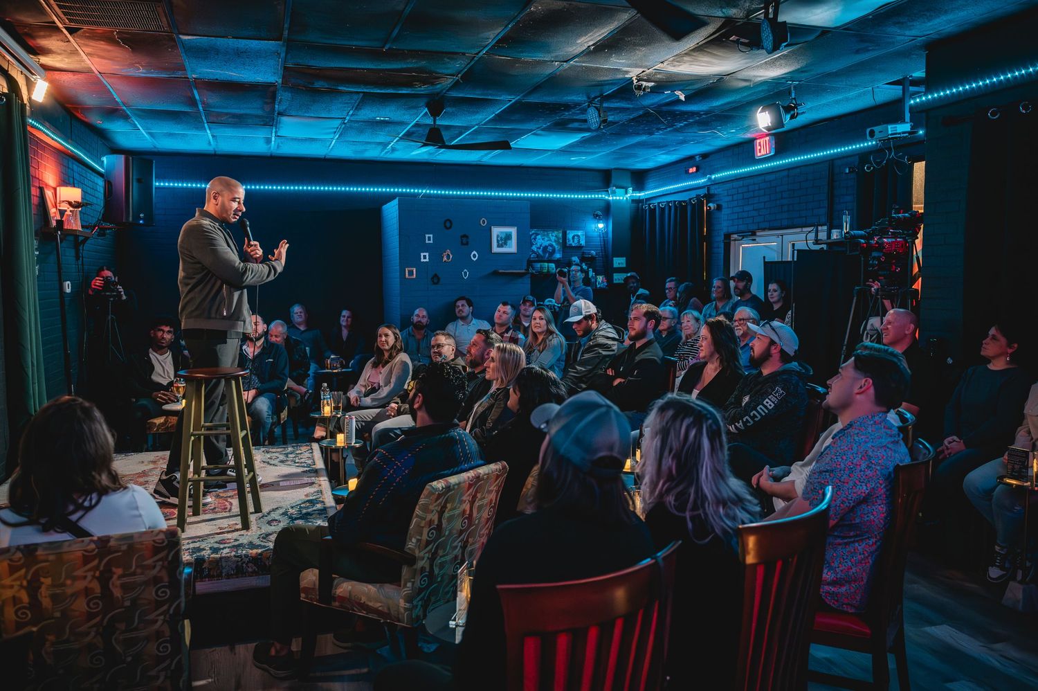 Comedian performs stand-up on stage in intimate dimly lit comedy club with engaged audience seated around candle-lit tables.