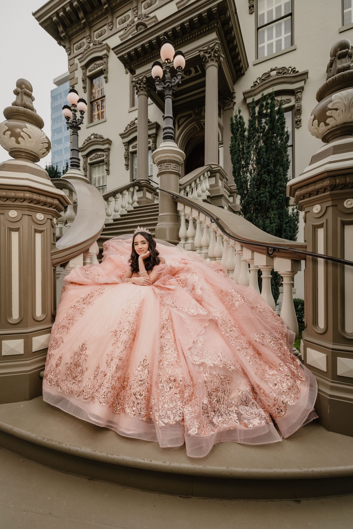 Young woman in a blue ball gown stands before ornate golden gates of a white castle-like building.