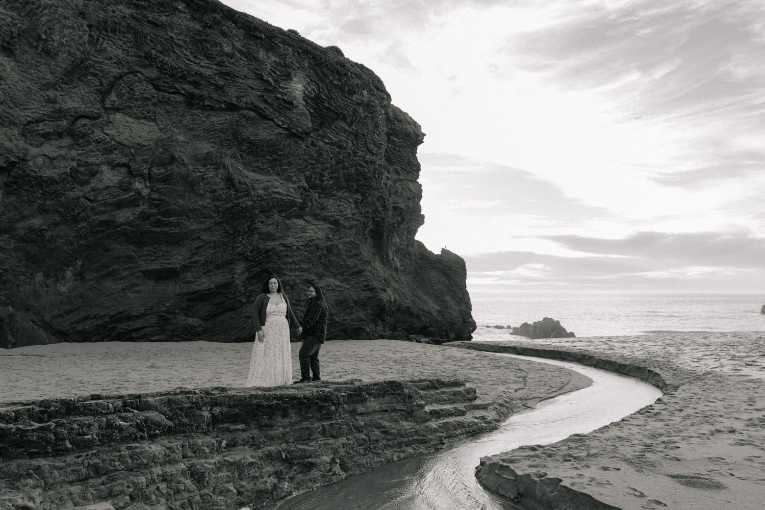 A couple in wedding attire stands on a dramatic black sand beach beside towering dark cliffs in a moody black and white photo.