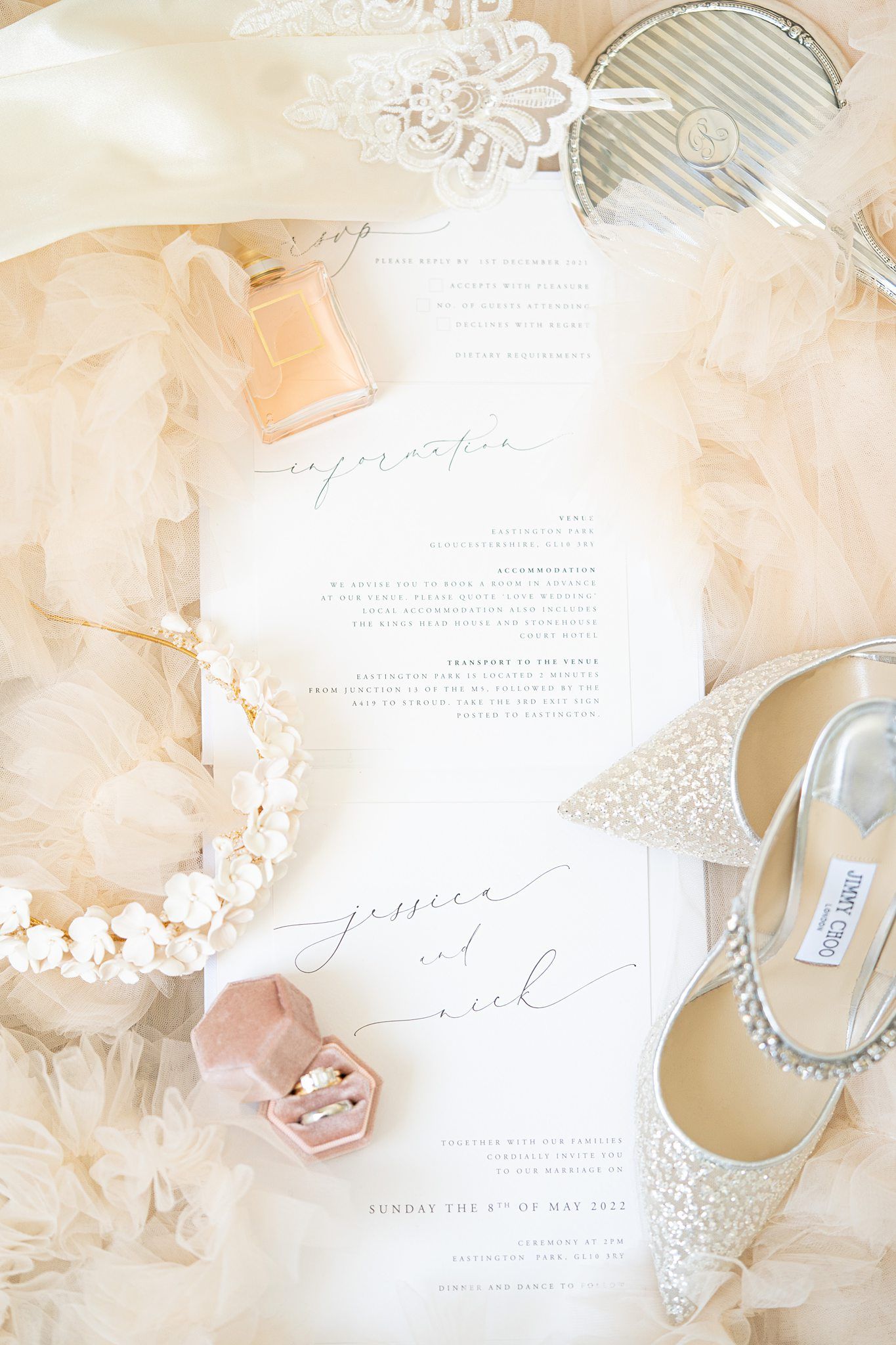 Elegant wedding flat lay with glittery heels, ring box, pearl bracelet, perfume, and calligraphy invitation on blush tulle.