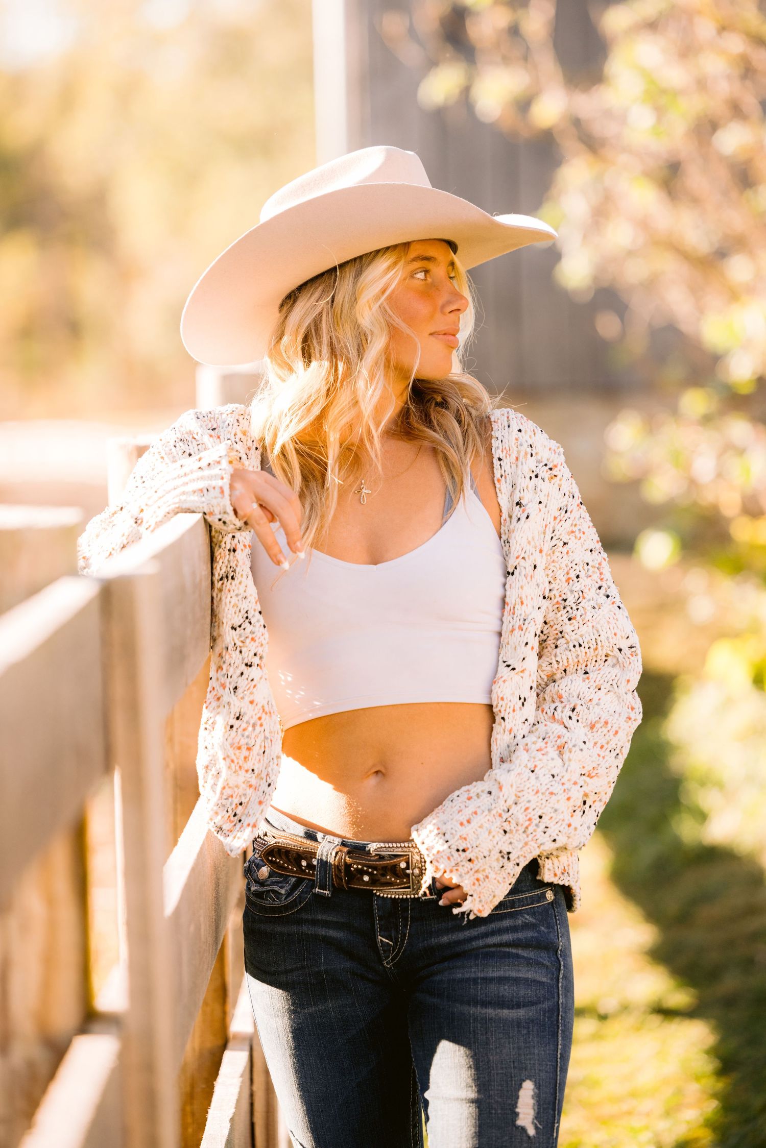 Western style outfit with white crop top, floral cardigan, distressed jeans and cowboy hat in golden sunlight.