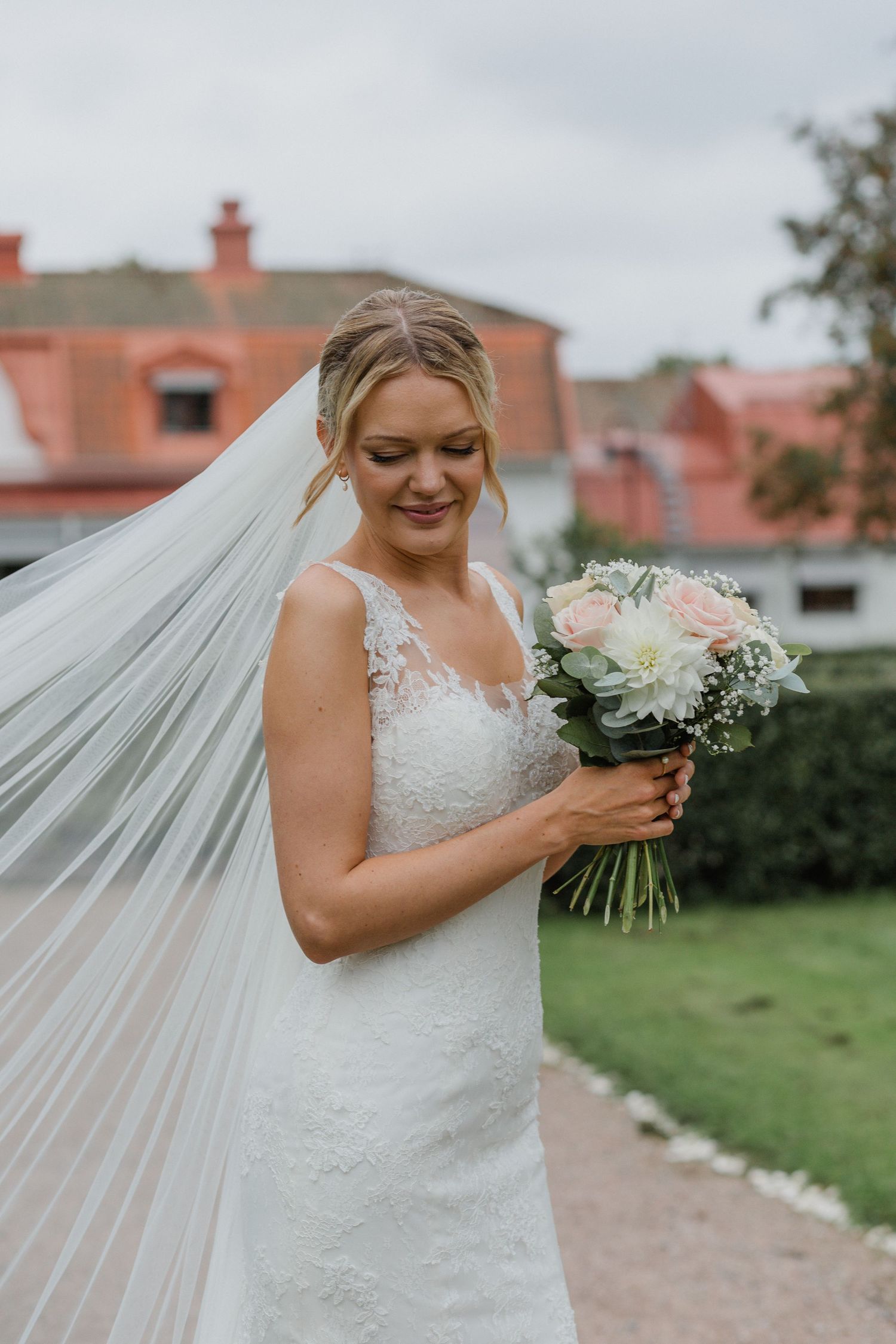 Bride in lace gown holds a bouquet of pink roses with veil flowing in the wind.