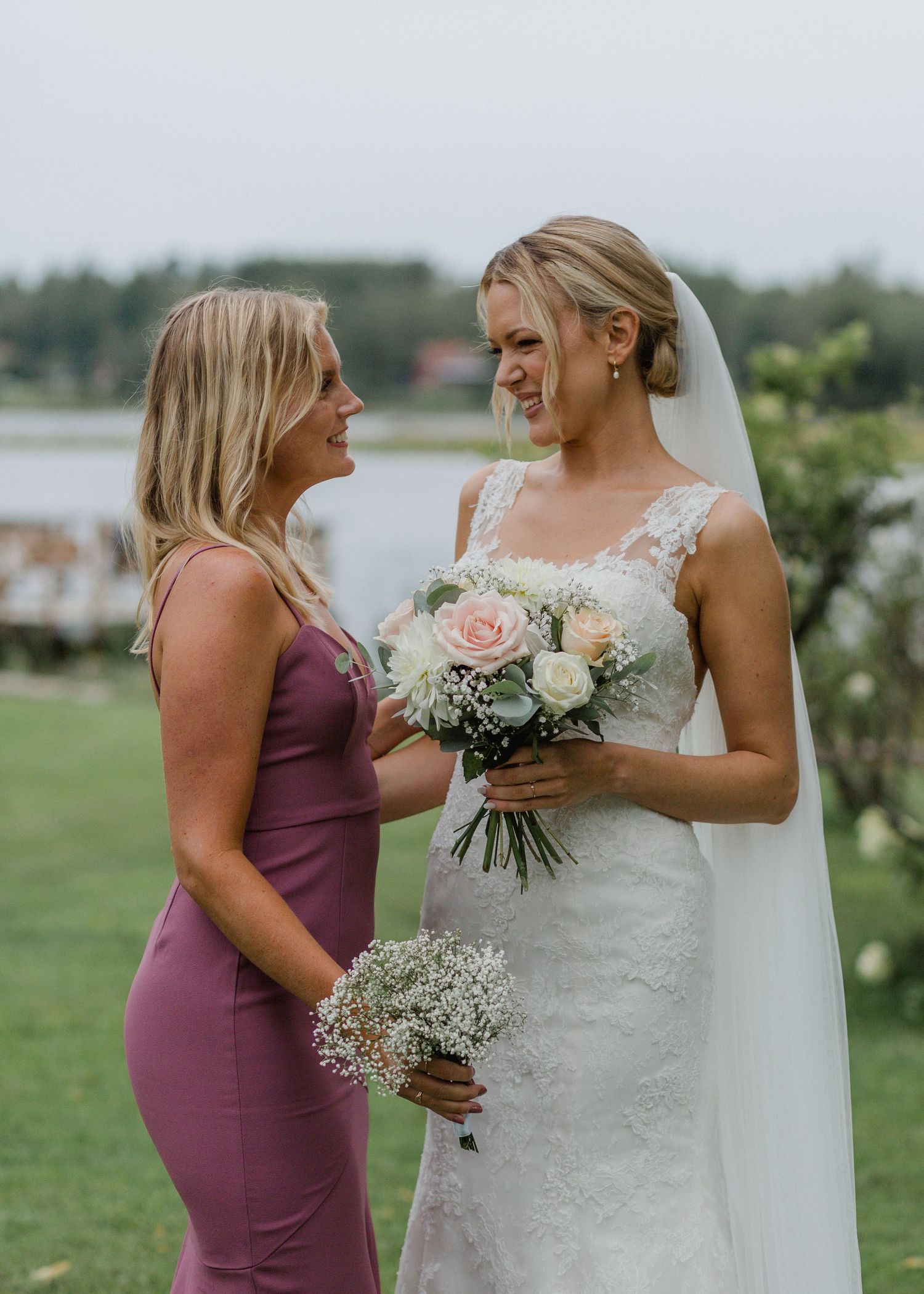 Bride in lace gown and bridesmaid in mauve dress share a joyful moment outdoors.