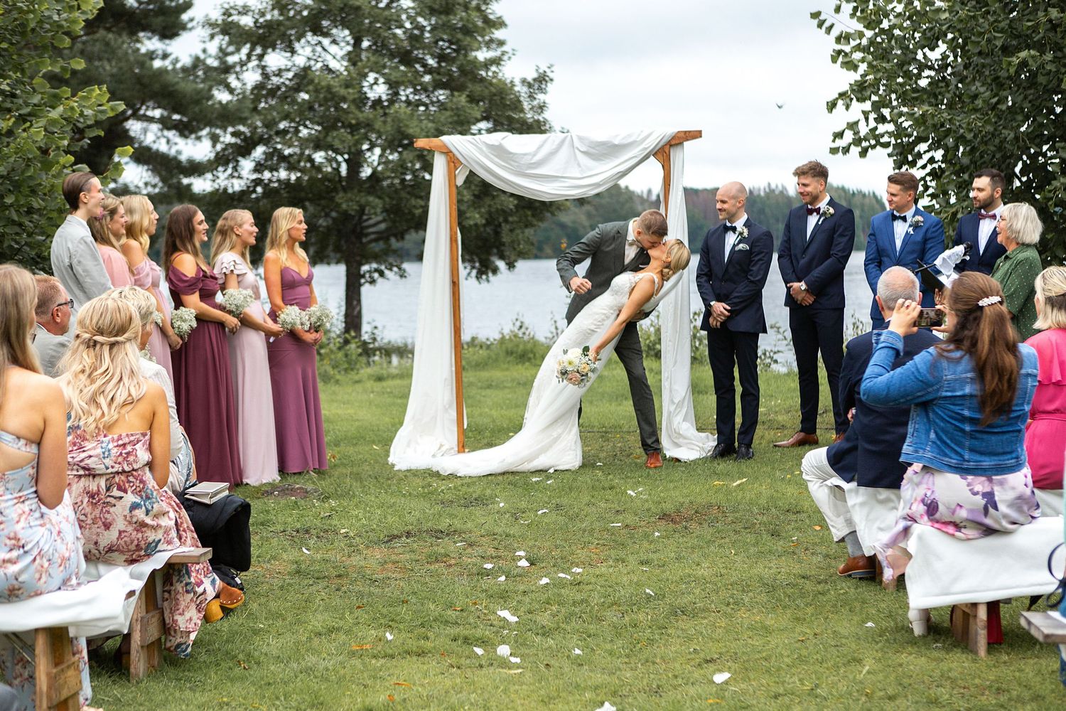 Couple shares a kiss at an outdoor wedding altar surrounded by guests and bridal party.